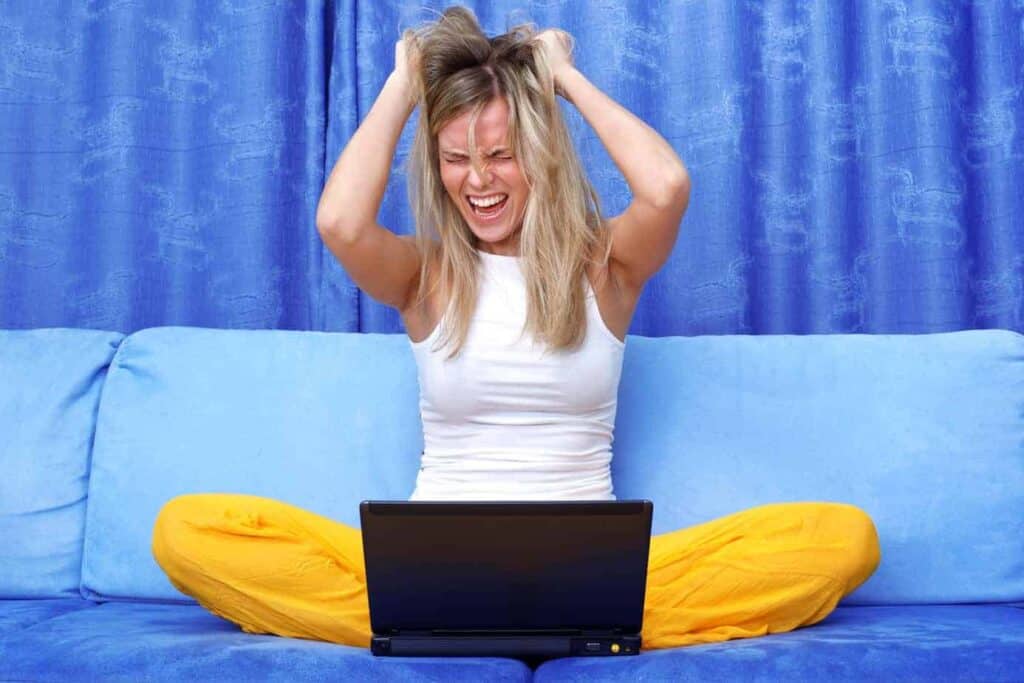 Alternative text: Woman looks stressed or frustrated while sitting on a blue sofa with a laptop, holding her head.