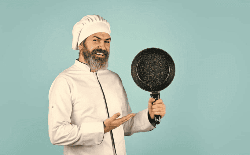 A smiling chef in a white uniform holding a black frying pan against a mint green background.