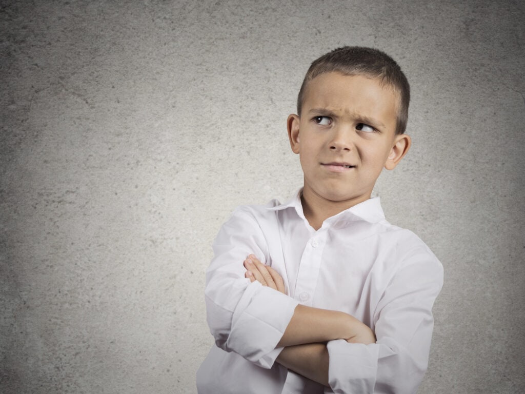 Child with a puzzled expression standing against a neutral textured wall.